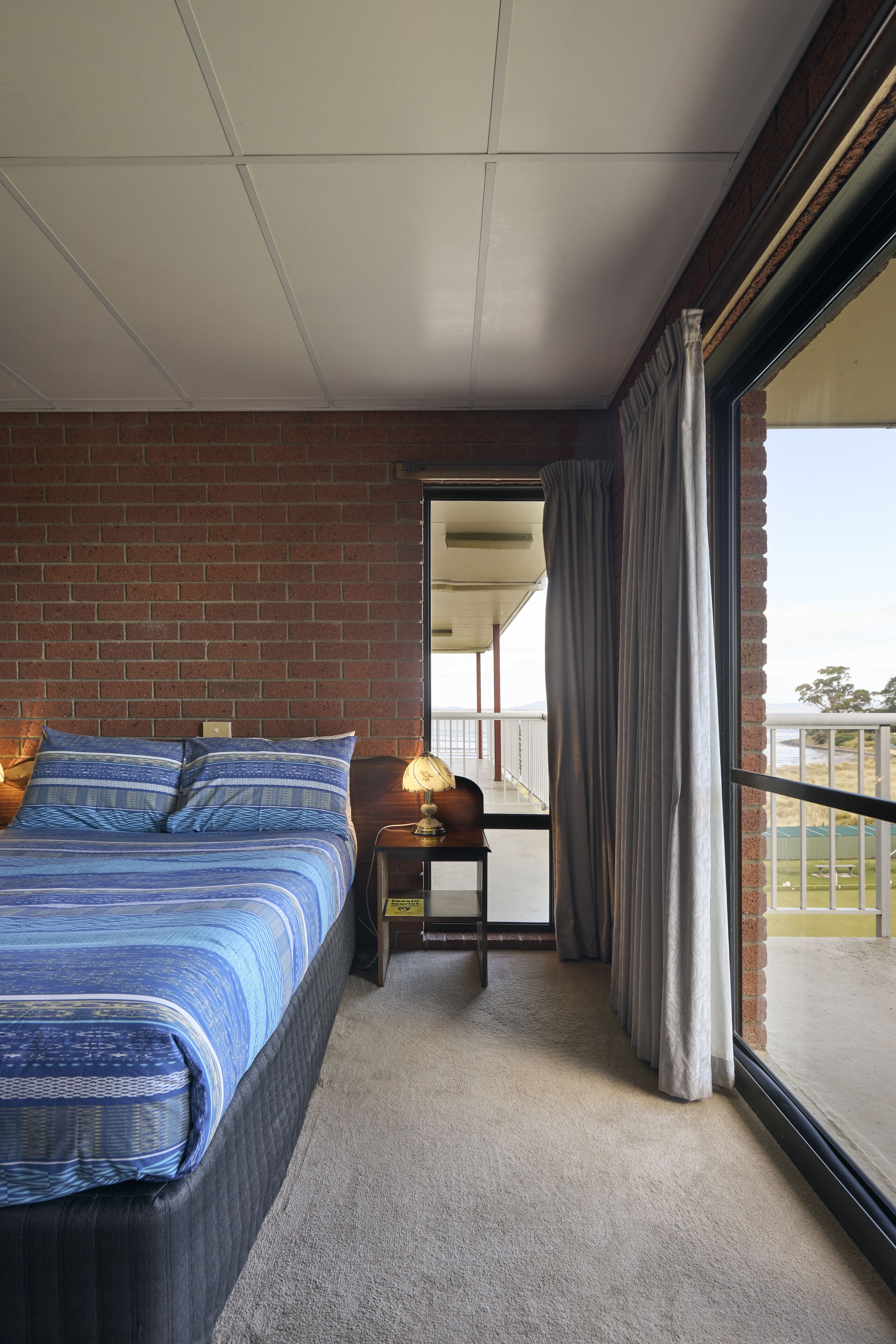 A bedroom with a brick wall, a large bed with blue bedding, a nightstand with a lamp, and a view of the outdoors through a sliding door.