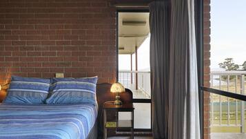 Un dormitorio con pared de ladrillo, una cama grande con ropa de cama azul, una mesita de noche con una lámpara y vistas al exterior por una puerta corredera.