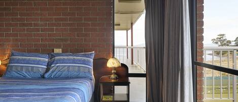 A bedroom with a brick wall, a large bed with blue bedding, a nightstand with a lamp, and a view of the outdoors through a sliding door.
