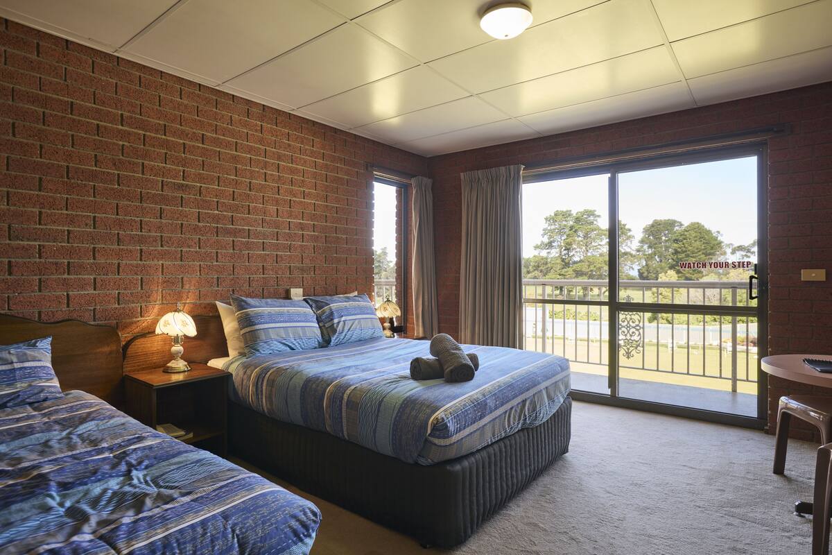 A hotel room with a brick wall, a large window with curtains, a bed with a striped bedspread, and a small table with a lamp.