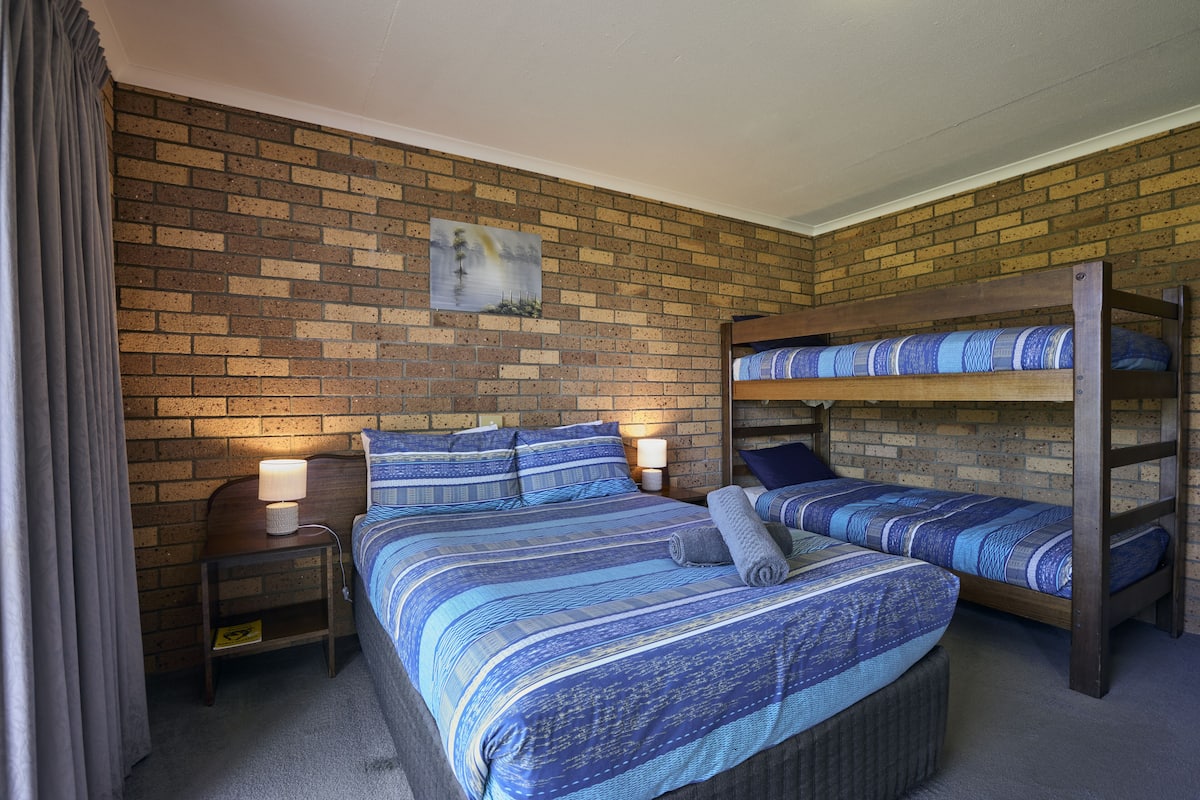 A room with a brick wall, a bed with blue bedding, a nightstand with a lamp, and a bunk bed.