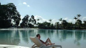 A person relaxing on a beach chair by a pool with a clear view of palm trees and a sandy shore.
