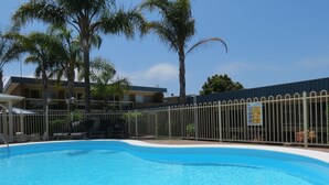 Ein Swimmingpool mit weißem Rand, umgeben von einem Metallzaun, Palmen und einem Gebäude mit einem Schild im Hintergrund.