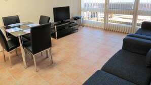 Ein Wohnzimmer mit Fernseher, Esstisch mit Stühlen, einer Couch und einem großen Fenster mit Jalousien.