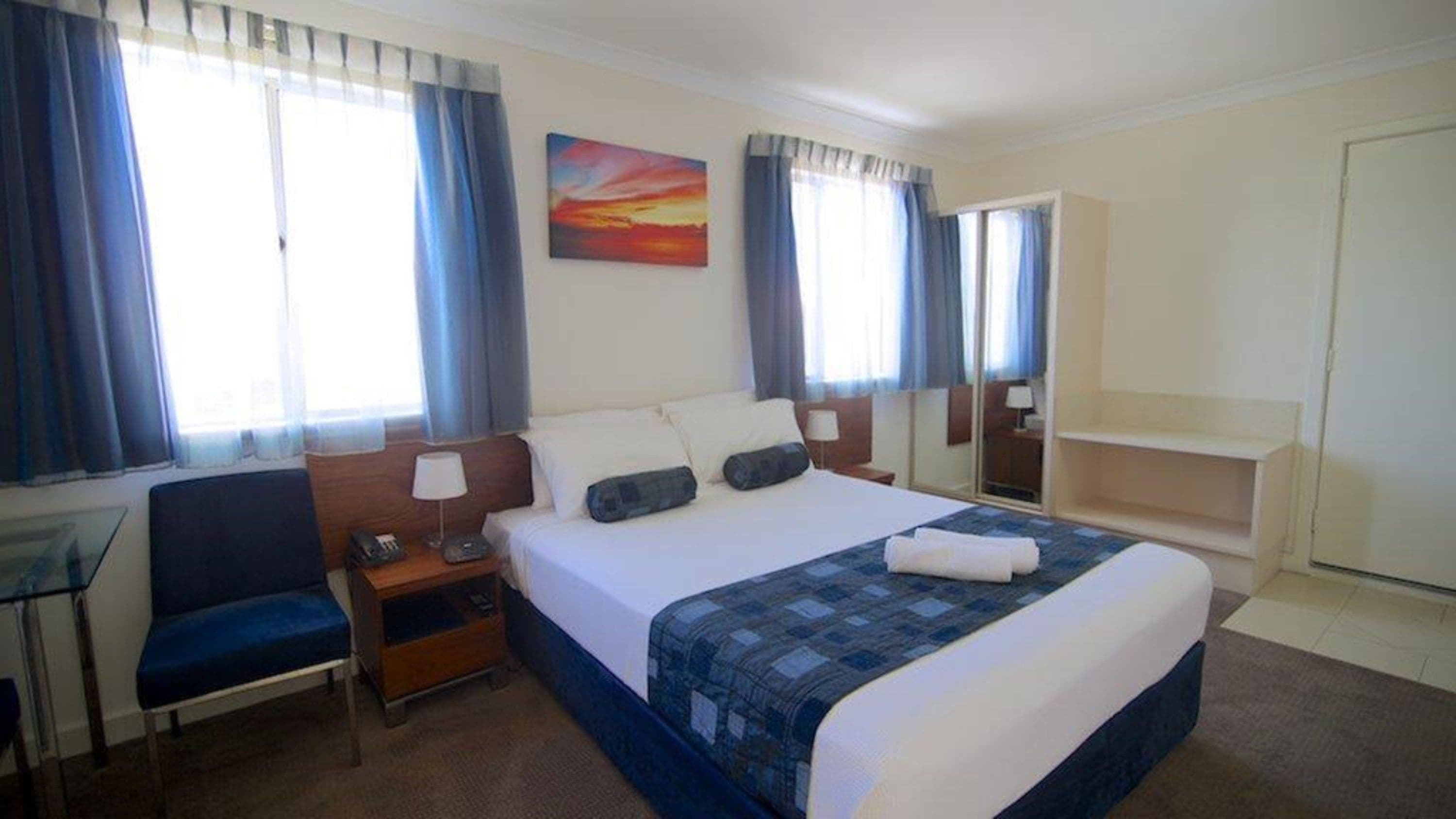 A hotel room with a bed, a desk, a chair, a nightstand, a painting, and a wardrobe.