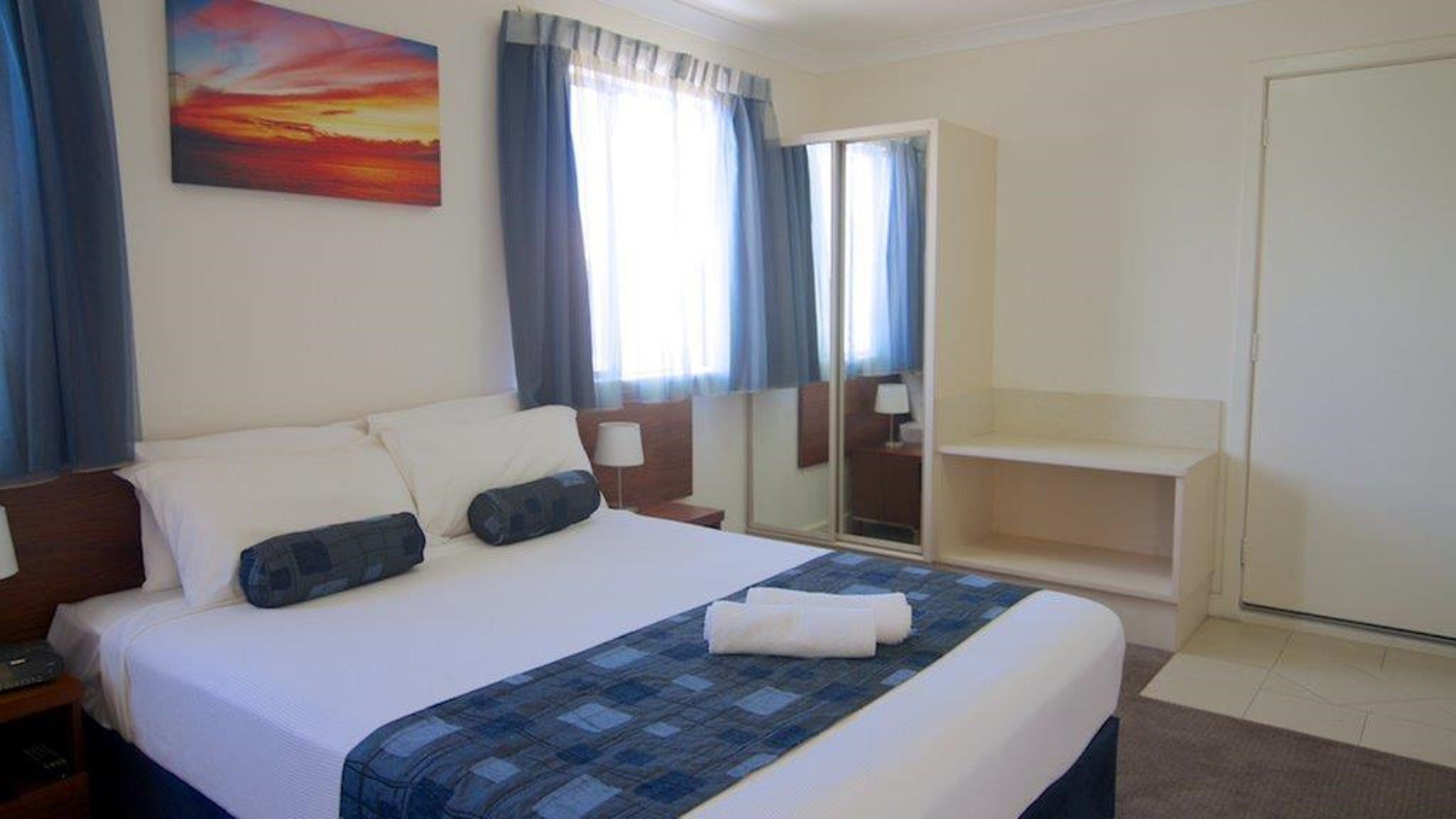 A hotel room with a bed, a nightstand, a desk, a mirror, and a window with curtains.