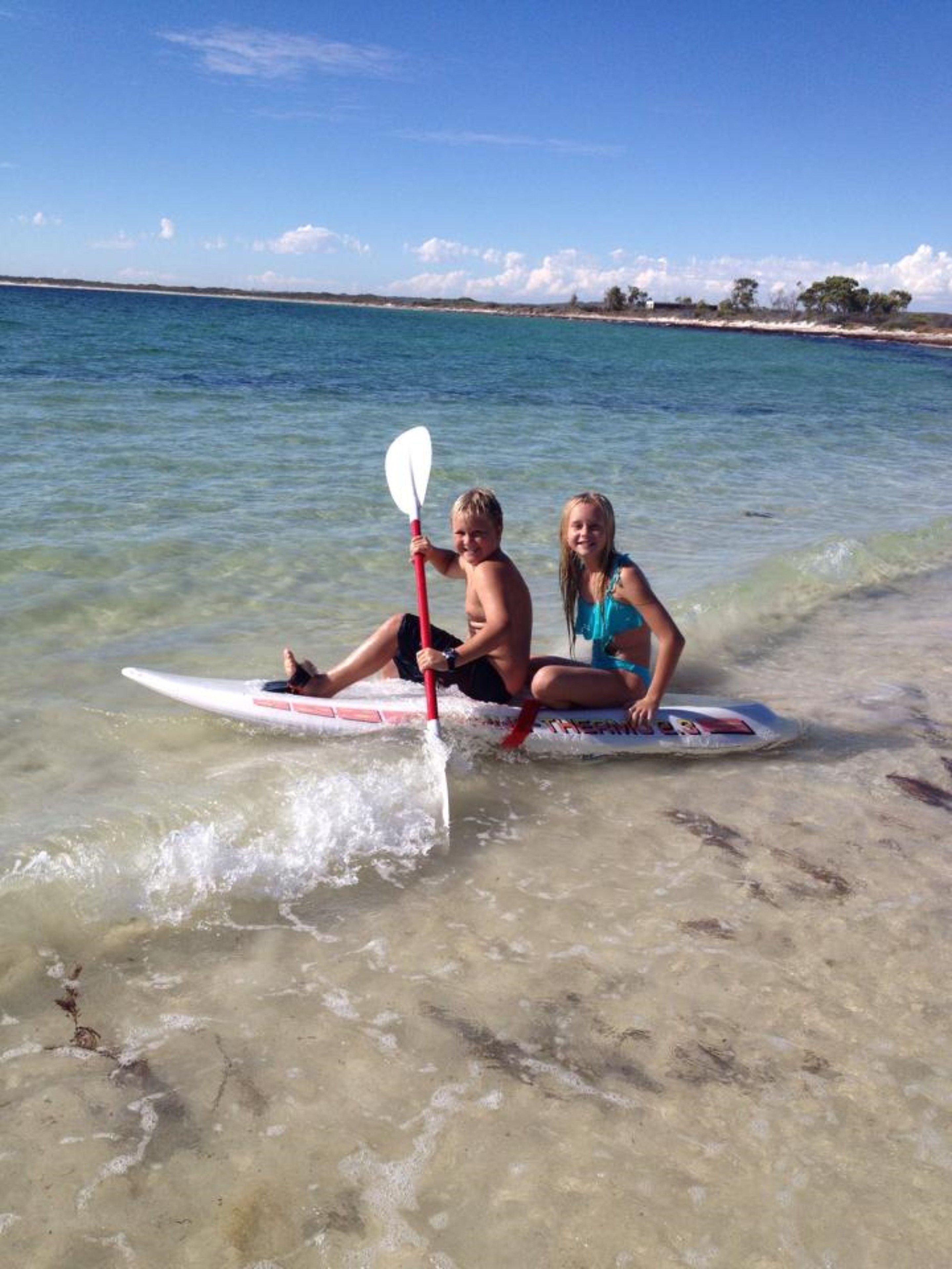 Two people sitting on a surfboard in shallow water with a clear view of the beach and ocean in the background.