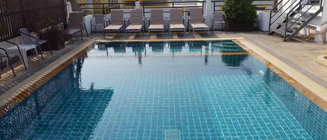 Piscina con suelo de baldosas y zona de estar con sillones de descanso y una escalera.