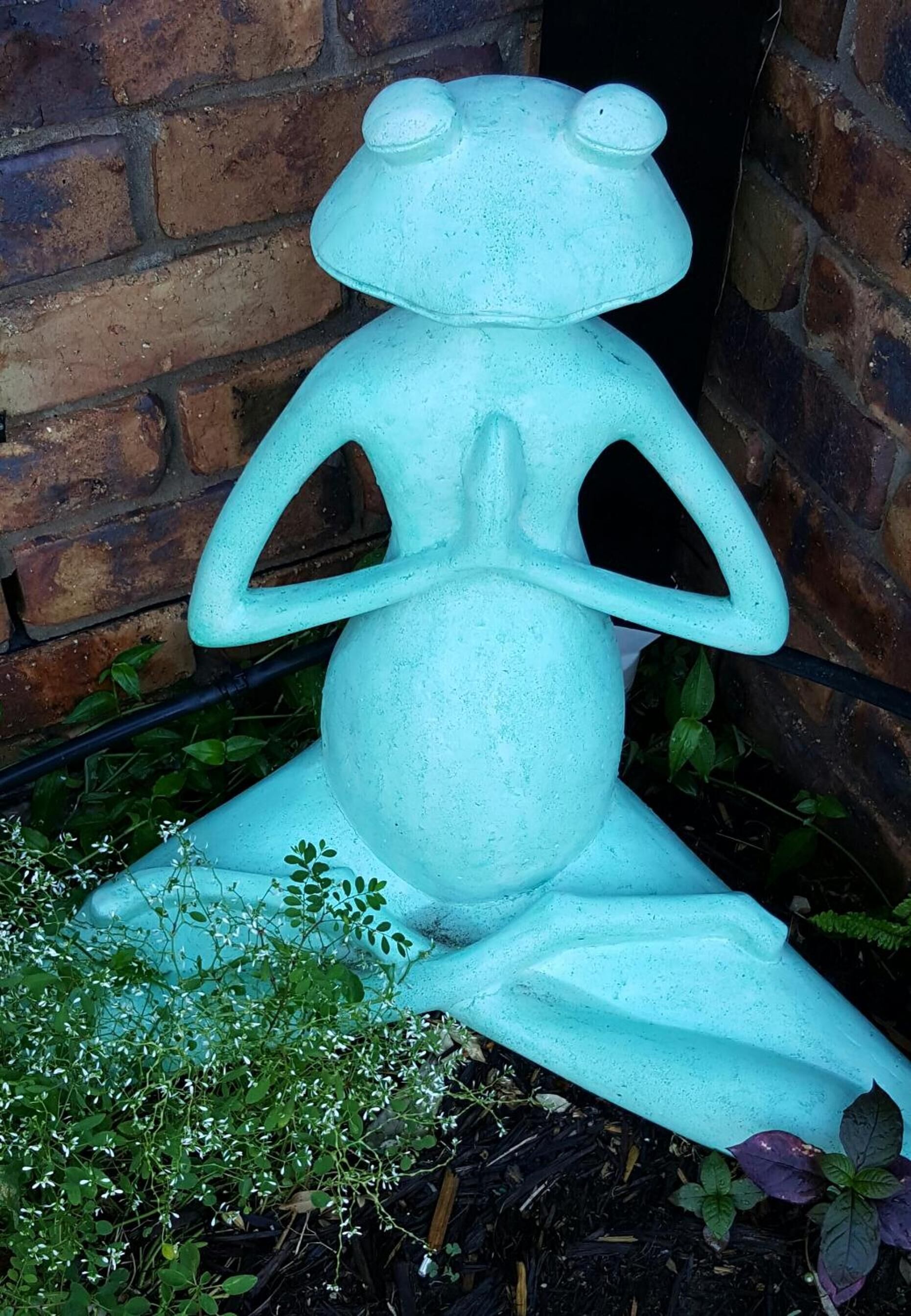 A sculpture of a frog in a seated position with its hands in a prayer gesture, placed on a brick surface with greenery around.