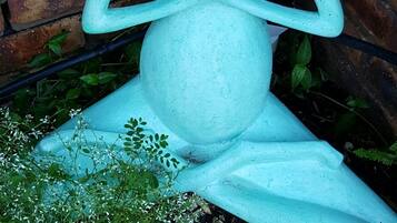 A sculpture of a frog in a seated position with its hands in a prayer gesture, placed on a brick surface with greenery around.