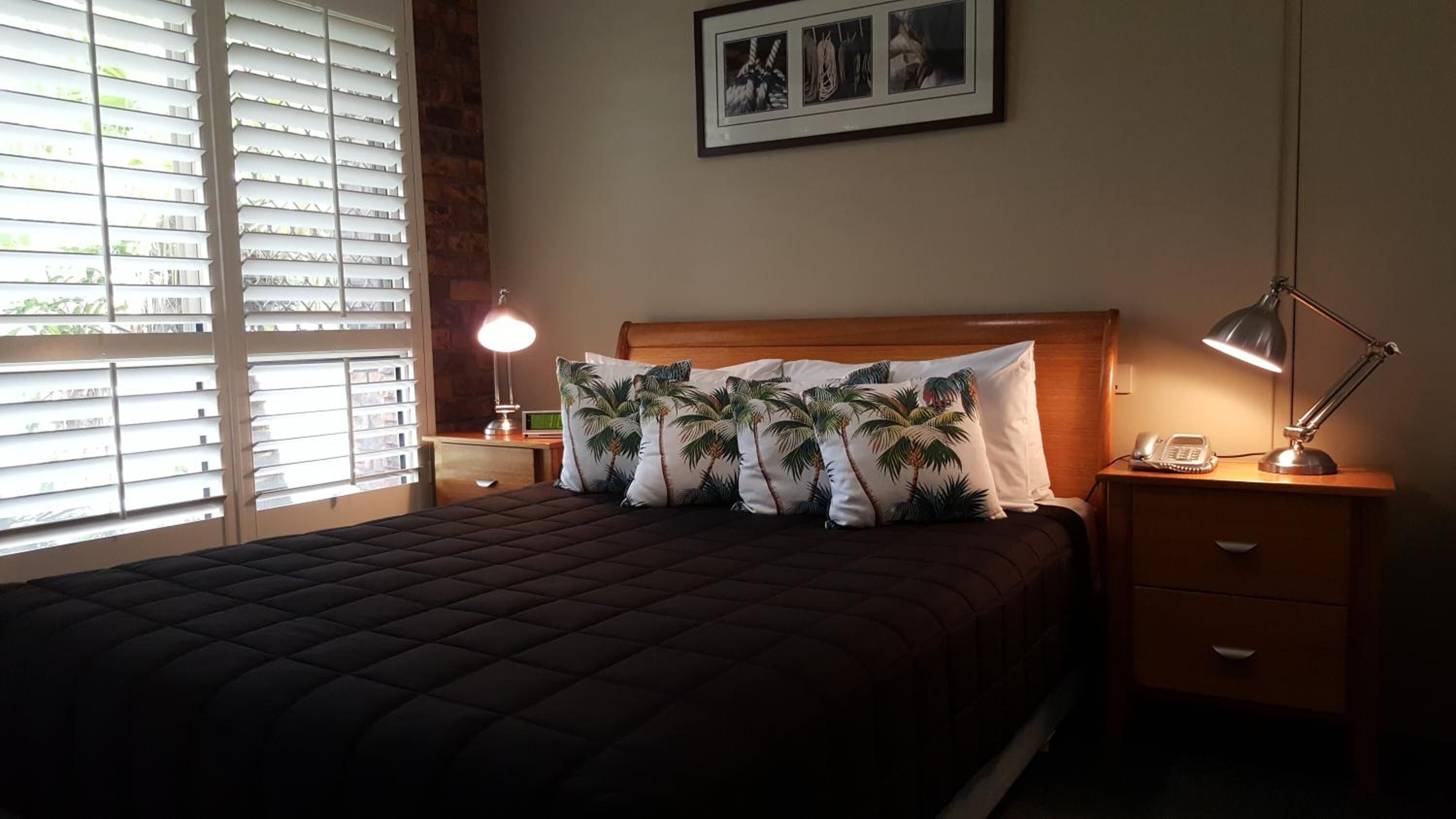 Un lit bien fait, recouvert d’une couverture sombre, avec une tête de lit en bois et des coussins à motifs de palmiers. On y trouve une lampe de chevet, une table de chevet avec un téléphone et un tableau encadré accroché au mur.