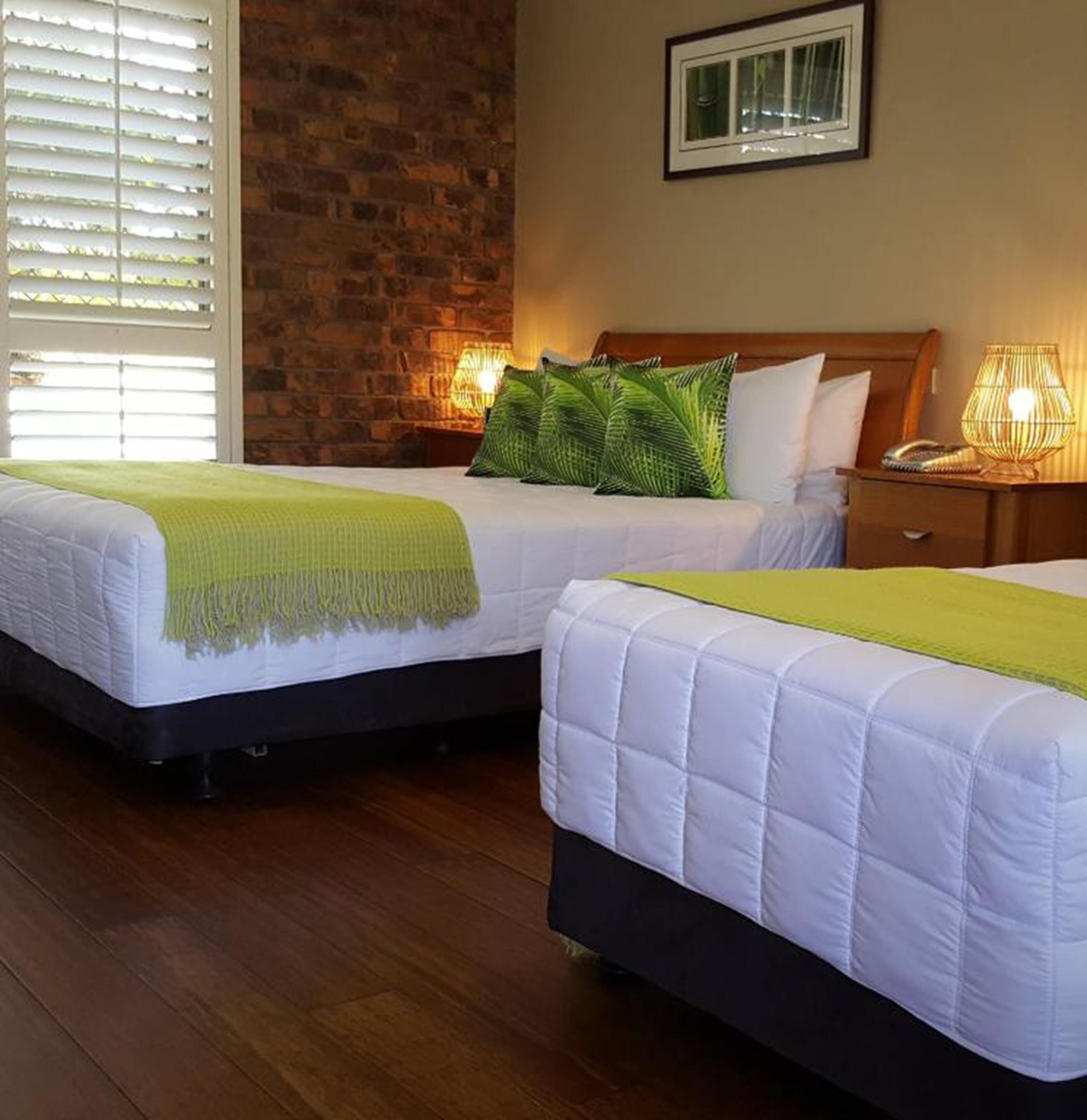 Two single beds with white bedding and green throw blankets, a brick wall, and a window with blinds.