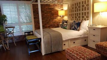 Ein Schlafzimmer mit Himmelbett, Backsteinwand, Fenster mit Jalousien, Nachttisch mit Lampe und einem Sessel.