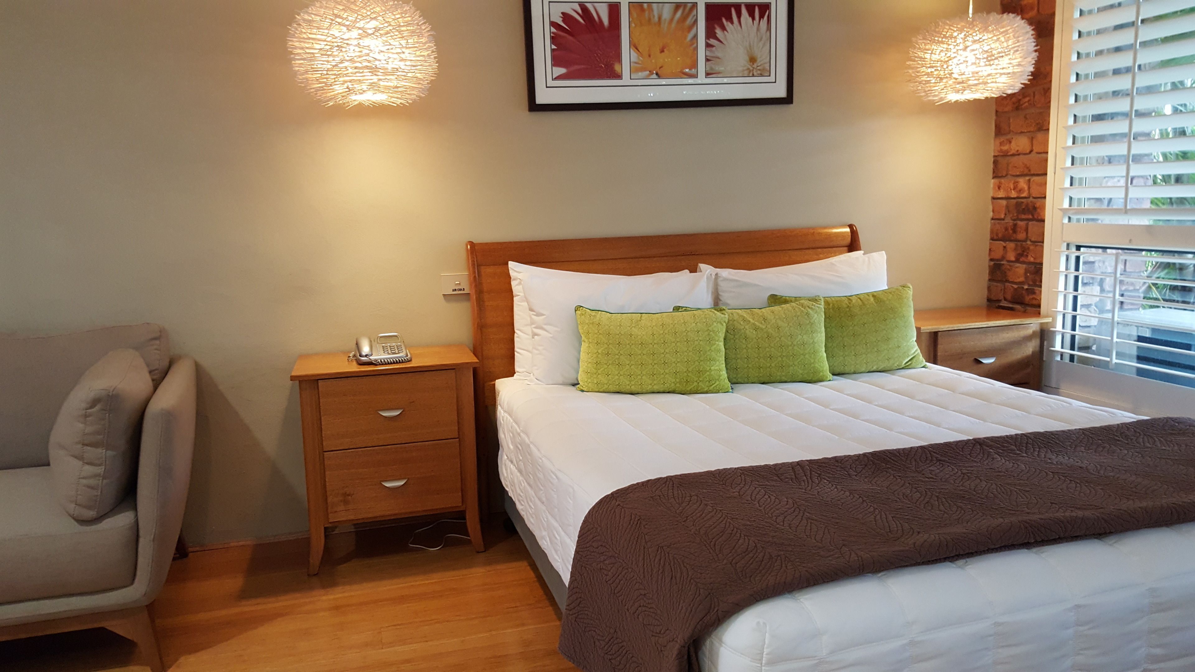 Un dormitorio ordenado con cabecera de madera, cama con almohadas blancas y verdes, mesita de noche con un teléfono y una silla.