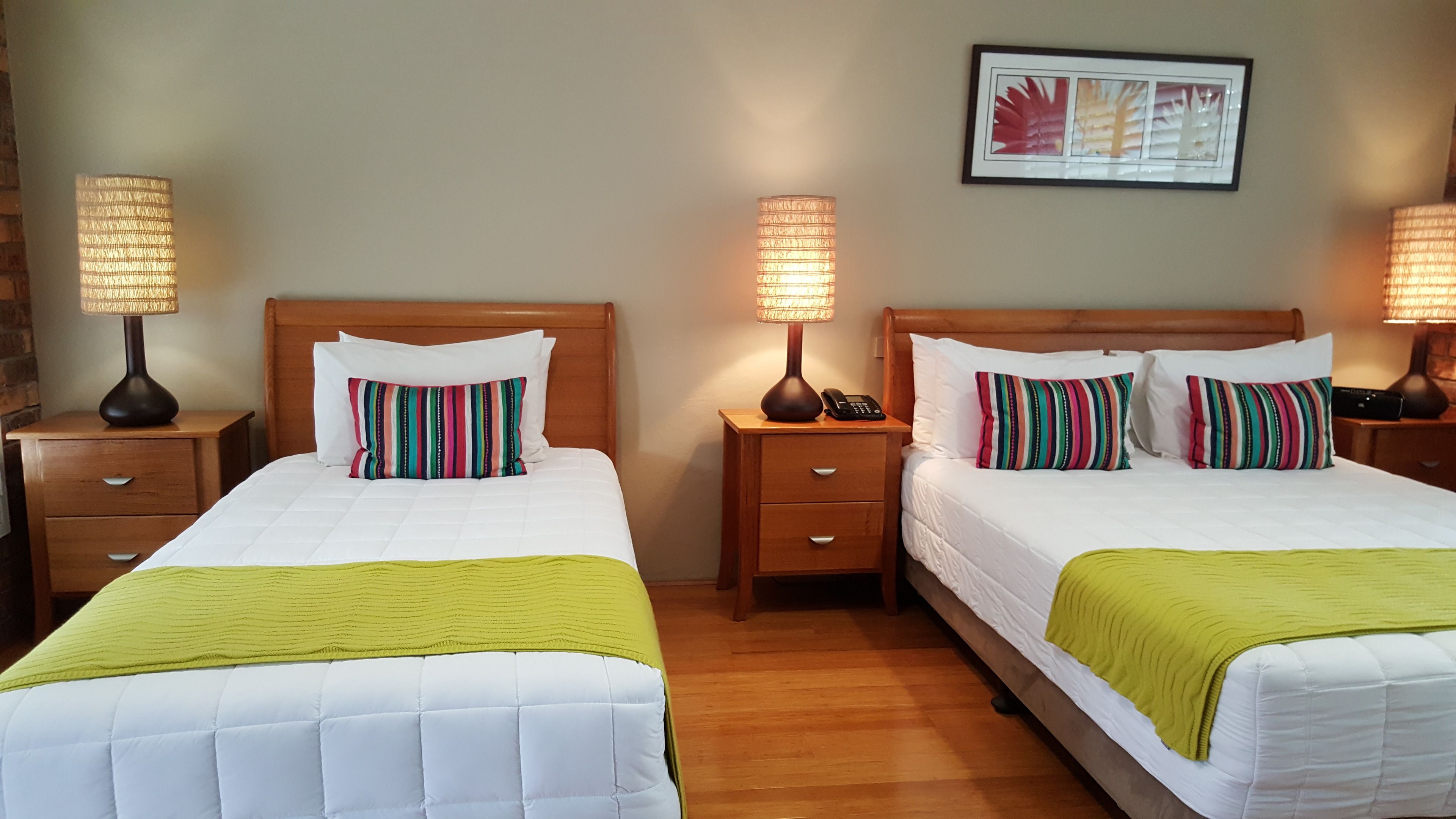 Habitación de hotel con dos camas, cada una con una colcha blanca y una manta verde. Se observan mesitas de noche de madera con lámparas y un cuadro en la pared.