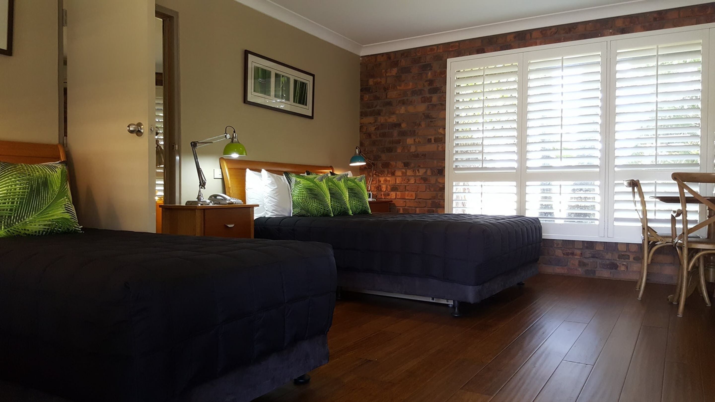 A hotel room with two beds, a brick accent wall, a large window with blinds, a desk with a chair, and a nightstand with a lamp.