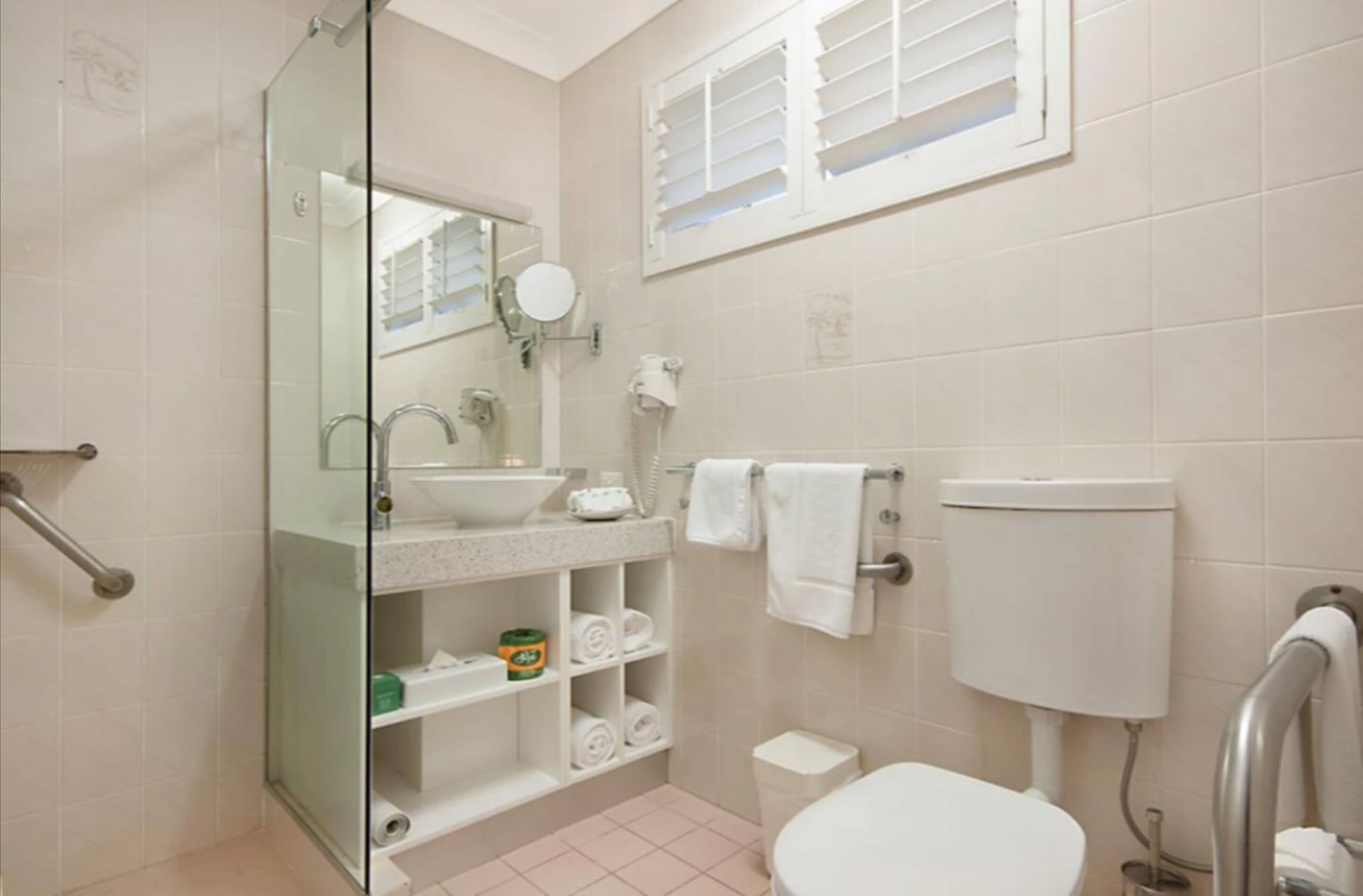 Een badkamer met een douchecabine van glas, een wastafel met spiegel, een toilet en een raam met luxaflex.