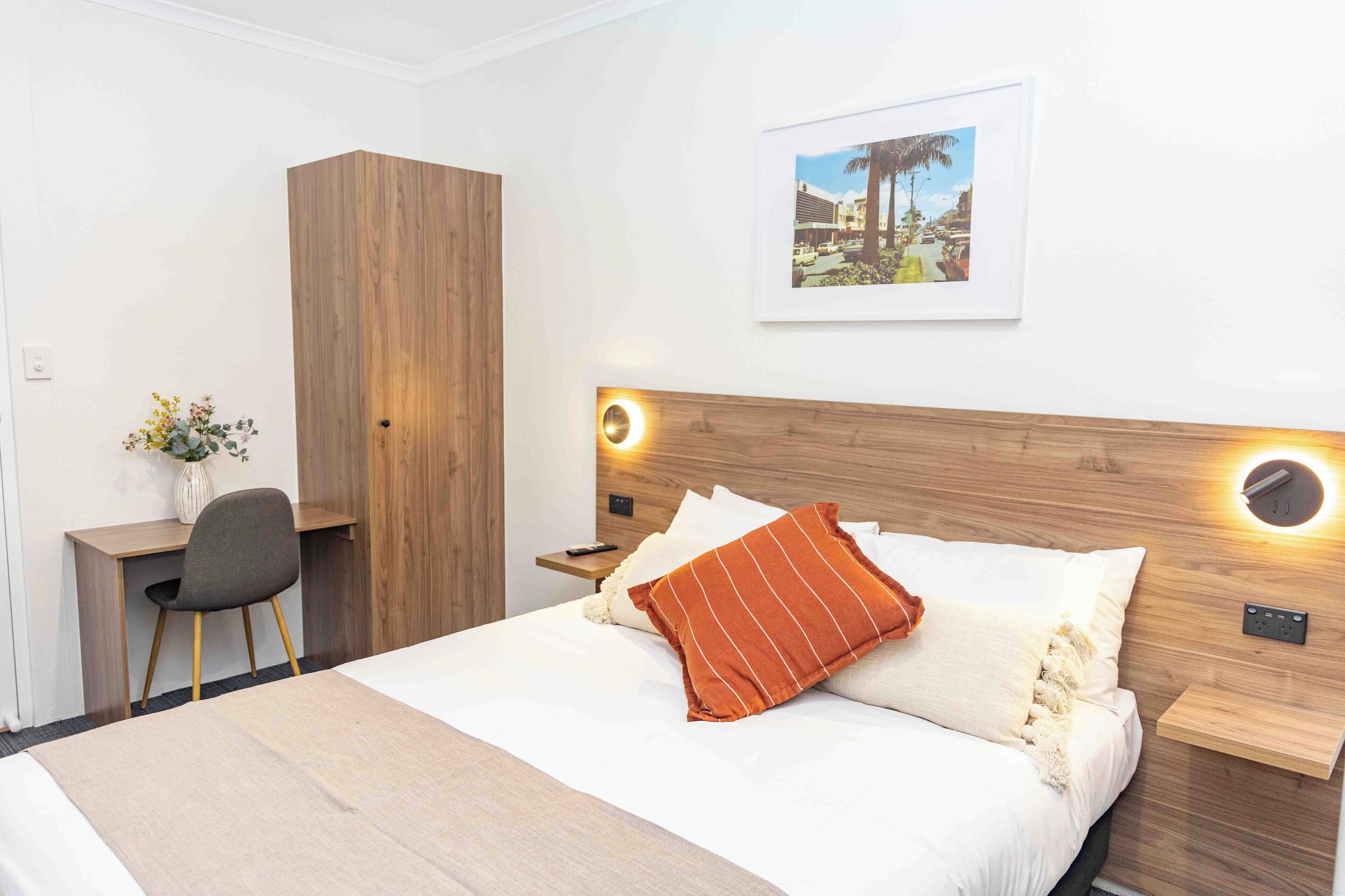 A hotel room with a bed, a desk, a chair, a wardrobe, and a wall-mounted picture.