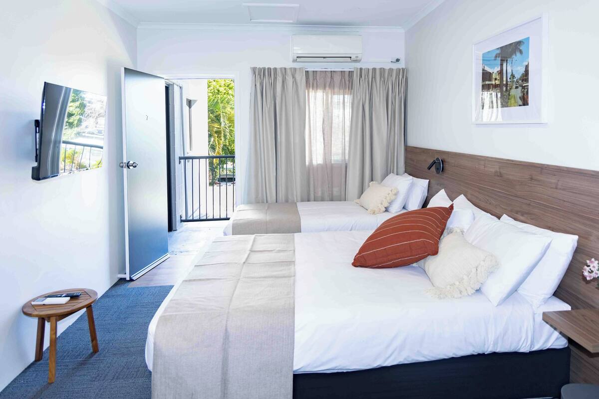 A hotel room with two beds, a TV, an air conditioning unit, and a balcony with curtains.