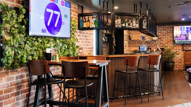 Een bar met een digitale scherm waarop een score van 77 staat, een bakstenen muur met beplanting en een toog met krukken.