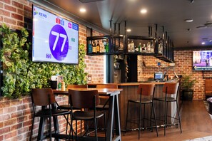 A bar area with a digital screen displaying a score of 77, a brick wall with greenery, and a bar counter with stools. - The Metropolitan Hotel (Mackay)