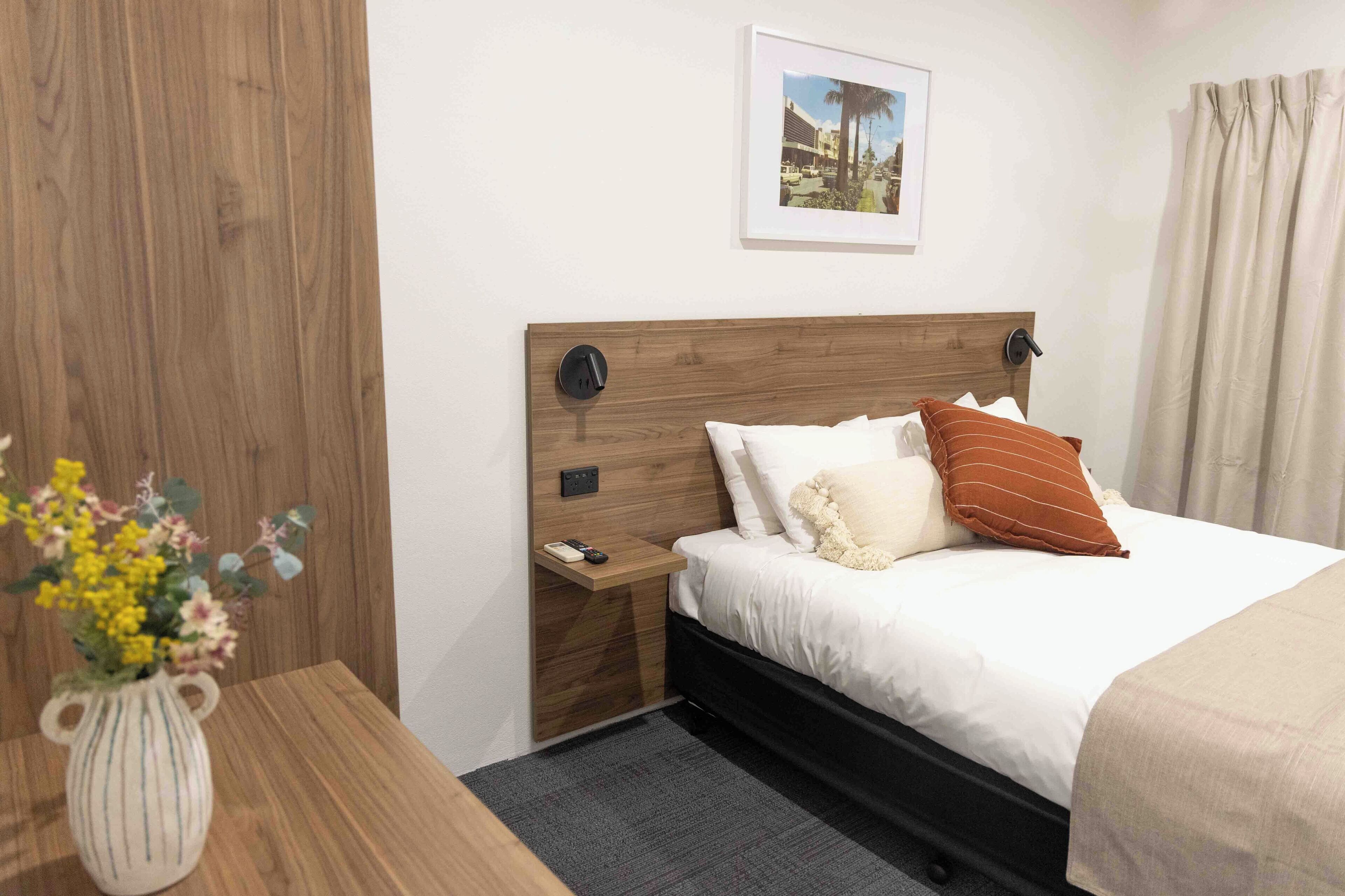 Une chambre d’hôtel avec une tête de lit en bois, une table de chevet ornée d’un vase de fleurs, un tableau encadré au mur et un lit avec une literie blanche et un oreiller marron.