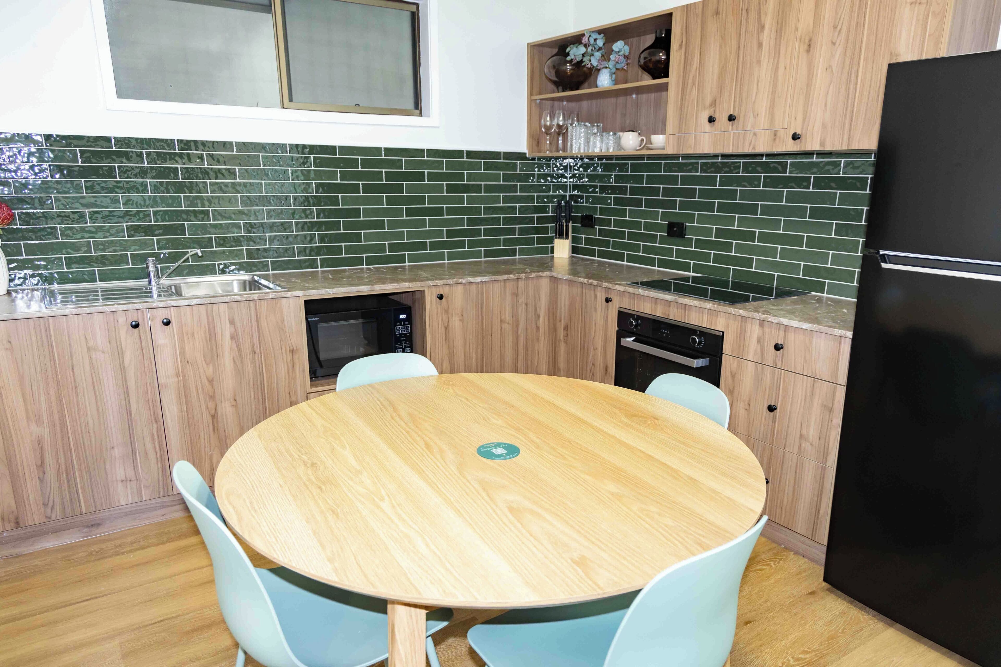 A kitchen with a round wooden table, light blue chairs, a black refrigerator, and wooden cabinets with green-tiled backsplash.