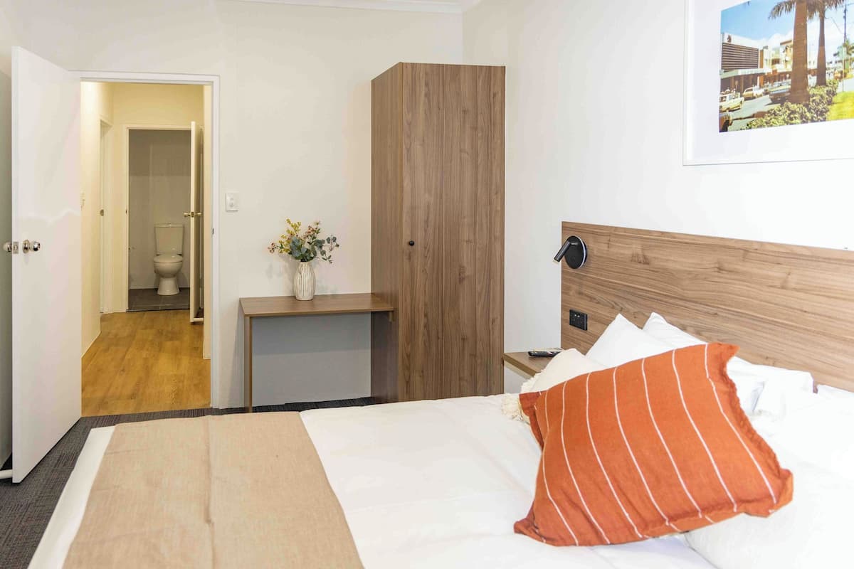 A hotel room with a bed, a wooden wardrobe, a bedside table with a vase, and a view of the outside.