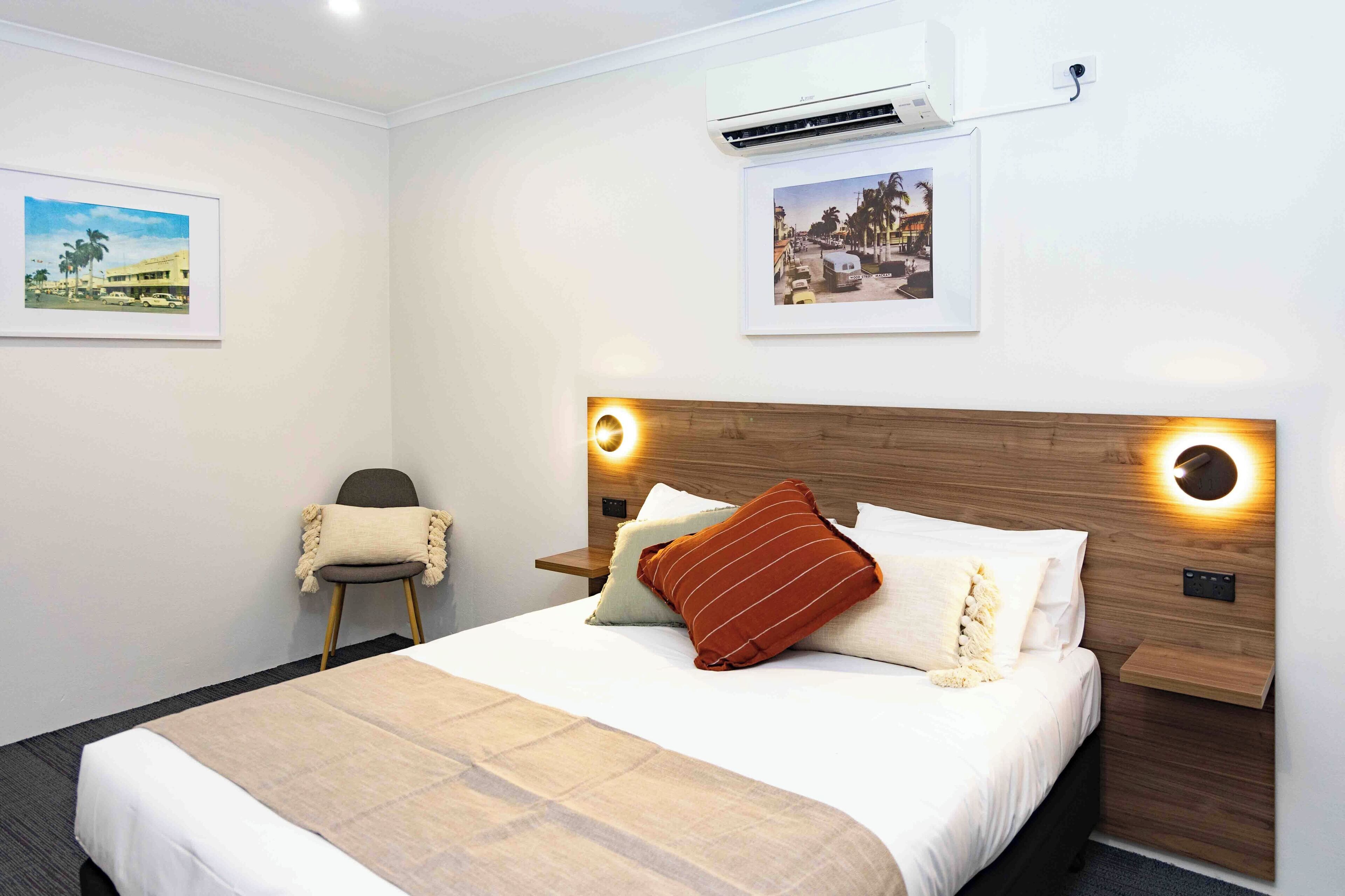 Una cama bien hecha con cabecera de madera, una mesita de noche y dos lámparas de lectura fijadas en la pared.