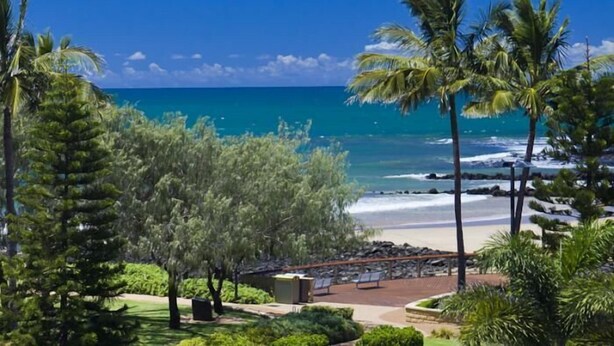 Vista a la playa con palmeras, una terraza de madera y un cielo azul despejado.
