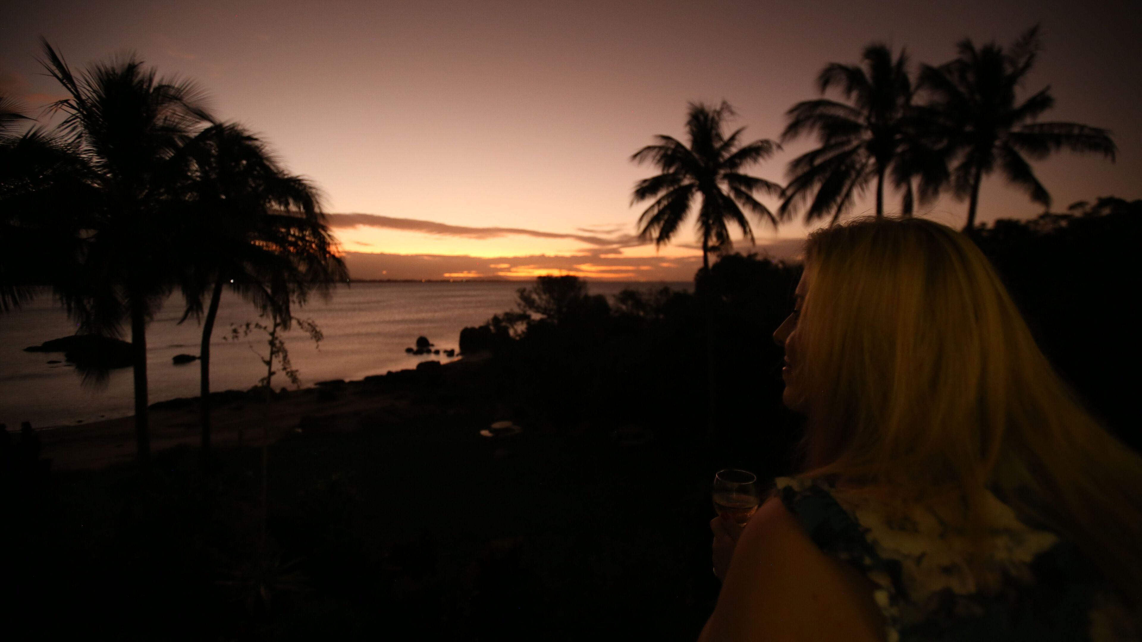 Eine Person blickt auf einen Sonnenuntergang mit Palmen und einem Strand.