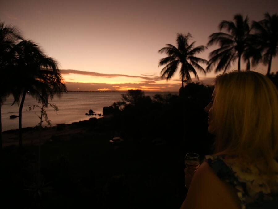 Eine Person blickt auf einen Sonnenuntergang mit Palmen und einem Strand.