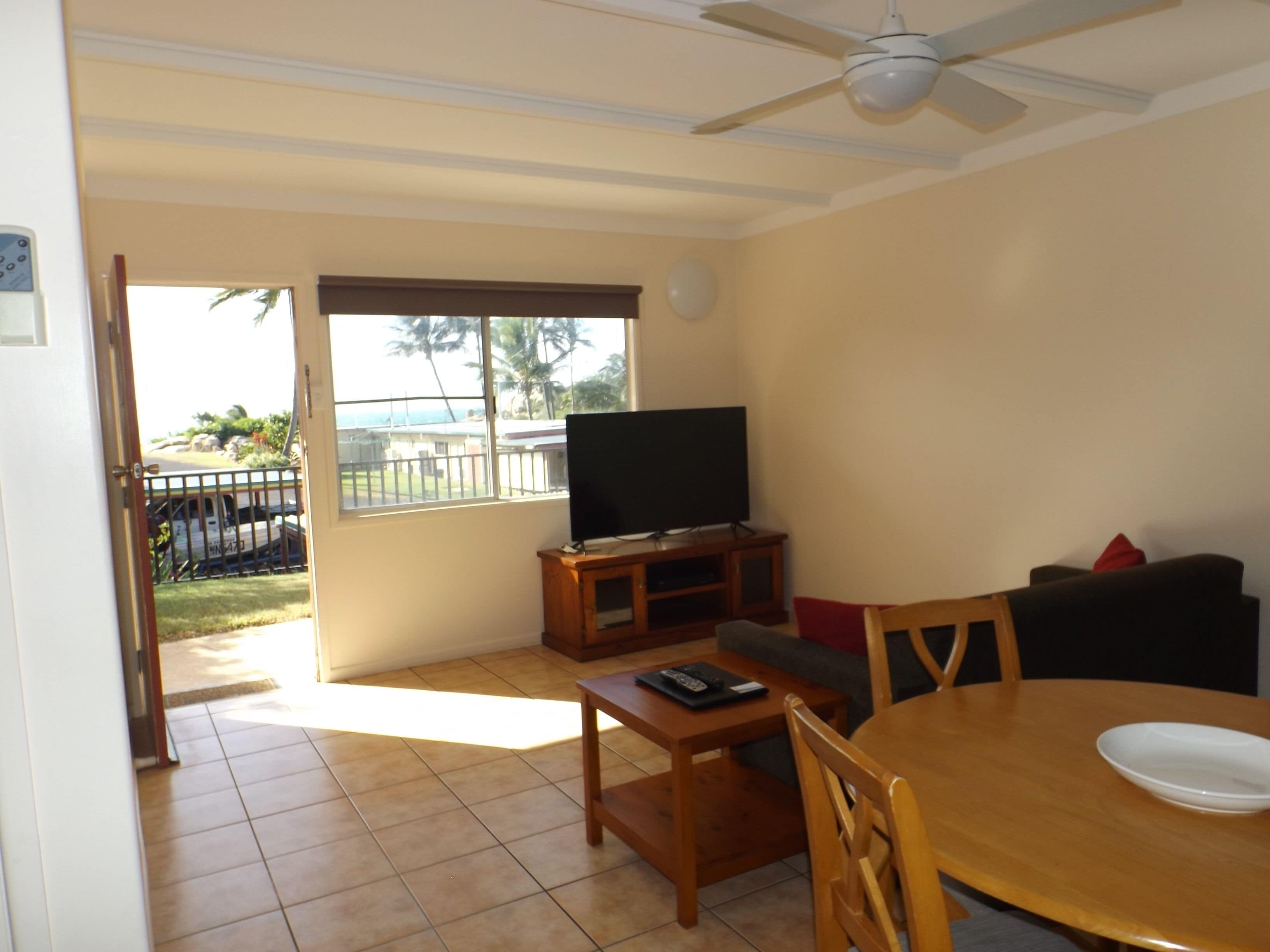 A living room with a TV on a wooden stand, a dining table with chairs, and a balcony with a view of palm trees.