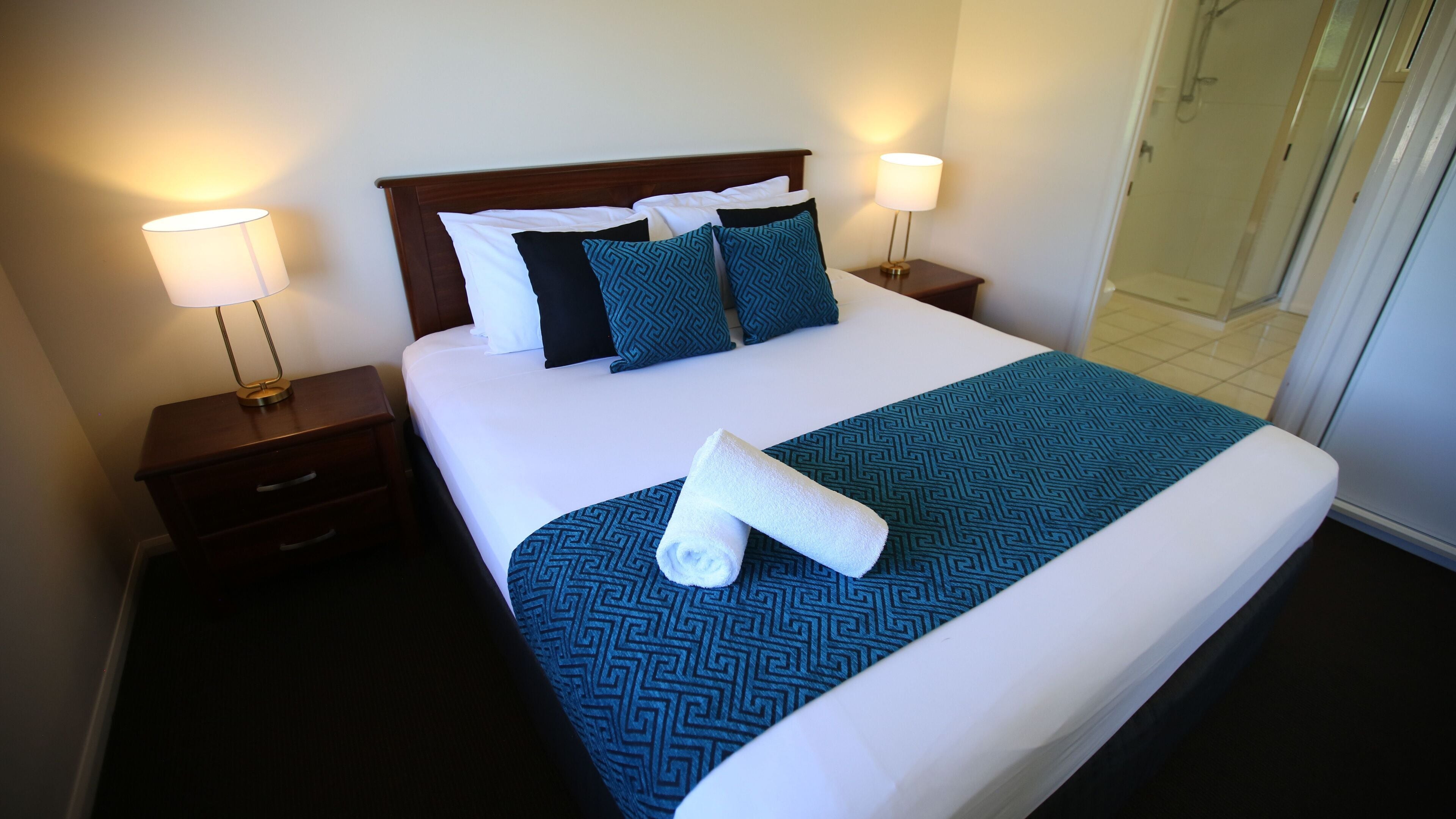 A neatly made bed with a patterned bedspread, two pillows, and a rolled towel on the bed. There are two bedside tables with lamps, one on each side of the bed.