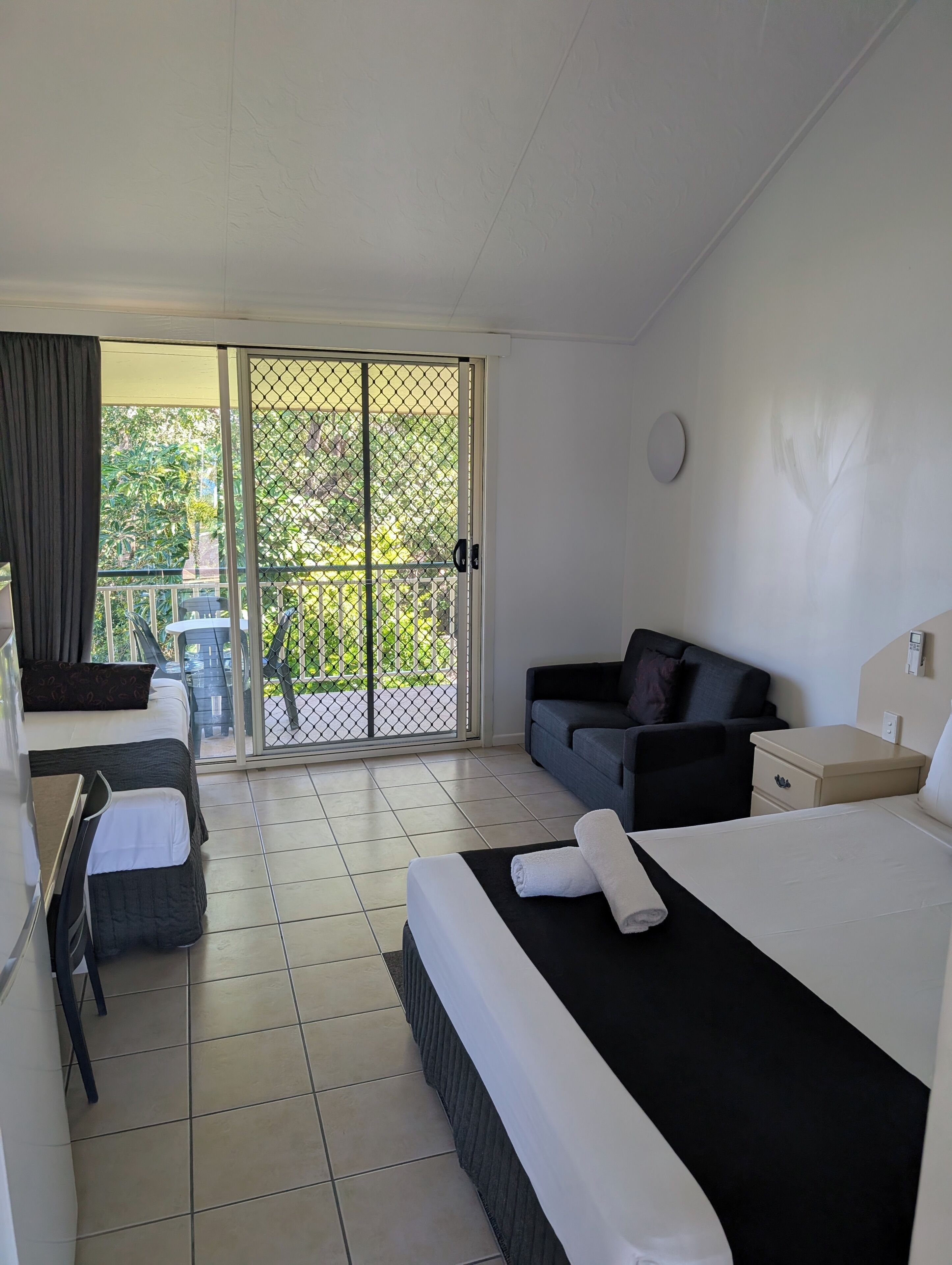 A hotel room with a bed, a sofa, a small table, and a balcony with a view of greenery.