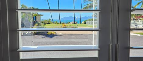 A view through a window with horizontal blinds, featuring a parking lot, a yellow golf cart, palm trees, and a clear blue sky.
