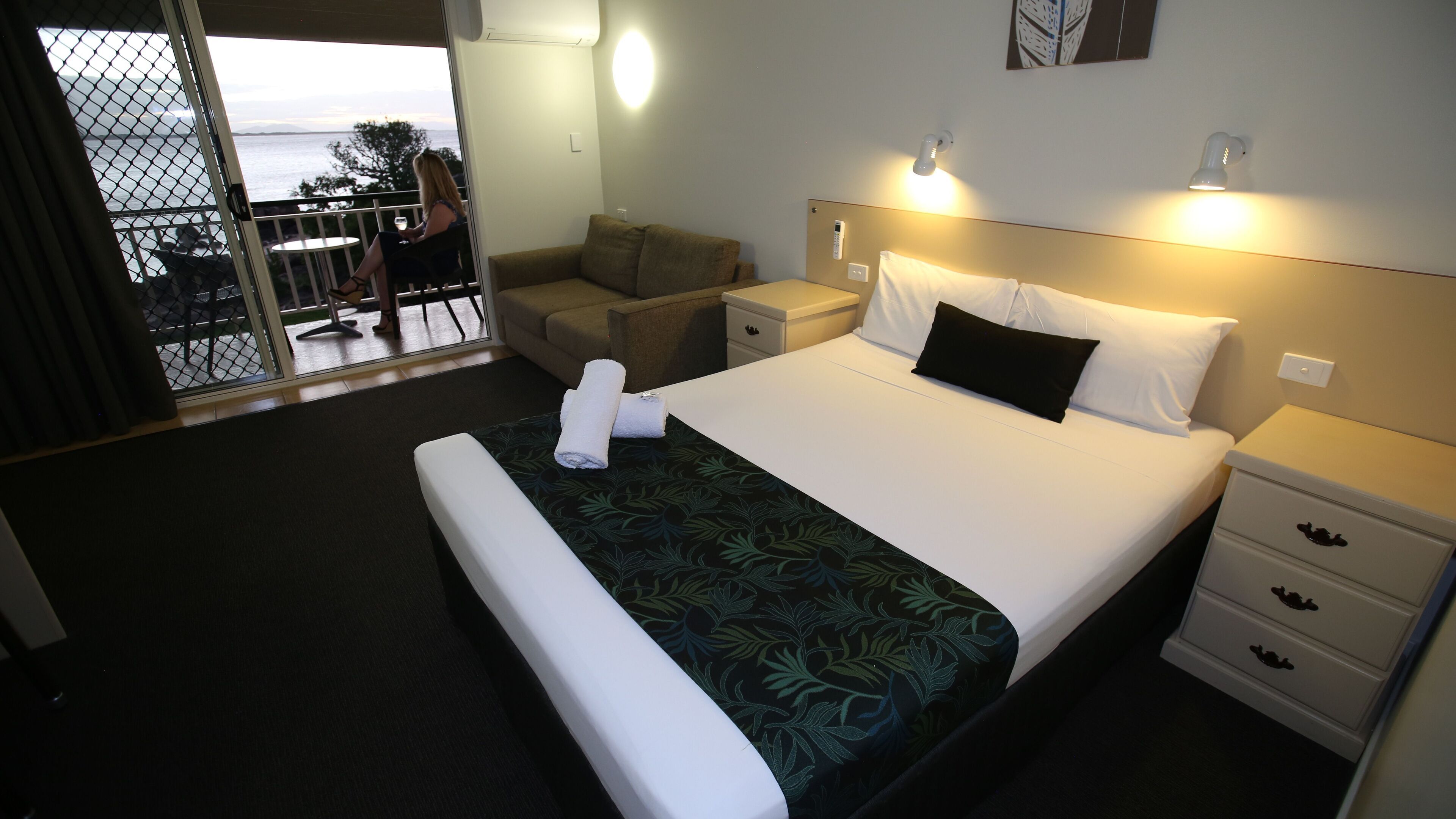 A hotel room with a bed, a nightstand, a chair, a small table, and a view of the sea through a balcony.