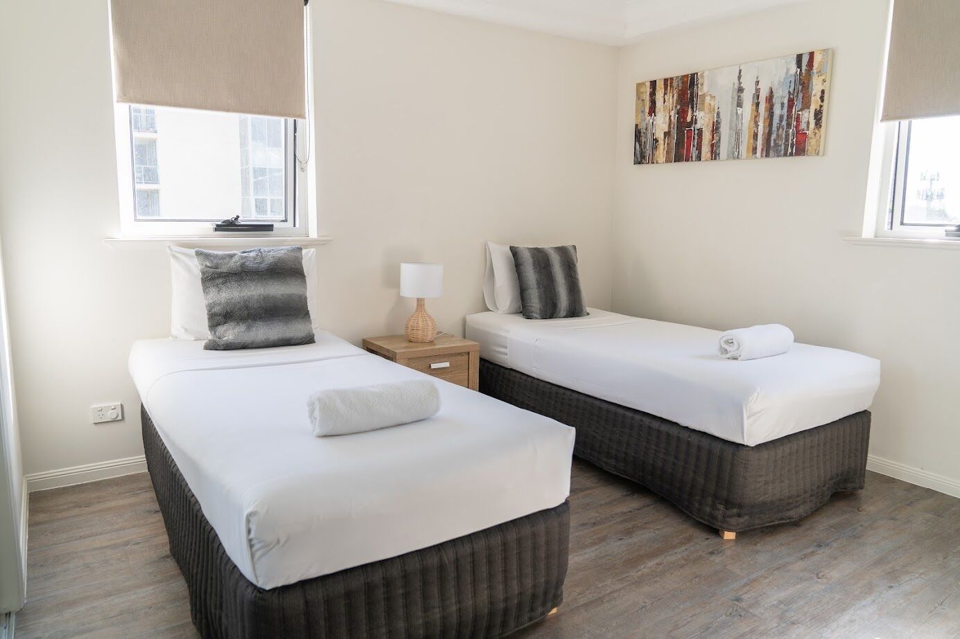 Habitación de hotel con dos camas individuales, una mesita de noche pequeña con lámpara, una obra de arte en la pared y una ventana con persiana.
