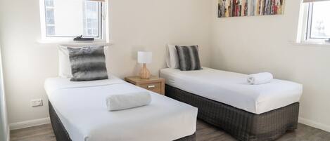 Habitación de hotel con dos camas individuales, una mesita de noche pequeña con lámpara, una obra de arte en la pared y una ventana con persiana.