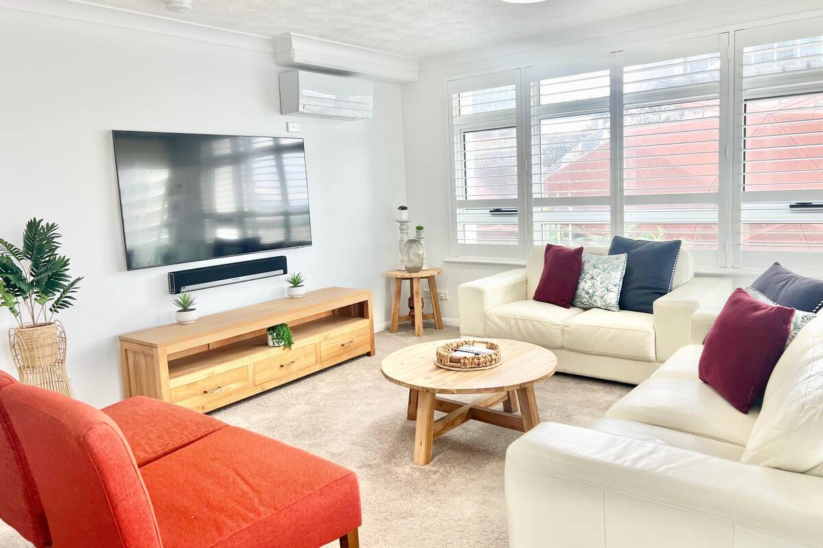 Un salon moderne avec un téléviseur à écran plat, un meuble de rangement en bois, un canapé blanc avec des coussins, un fauteuil rouge et une table basse.