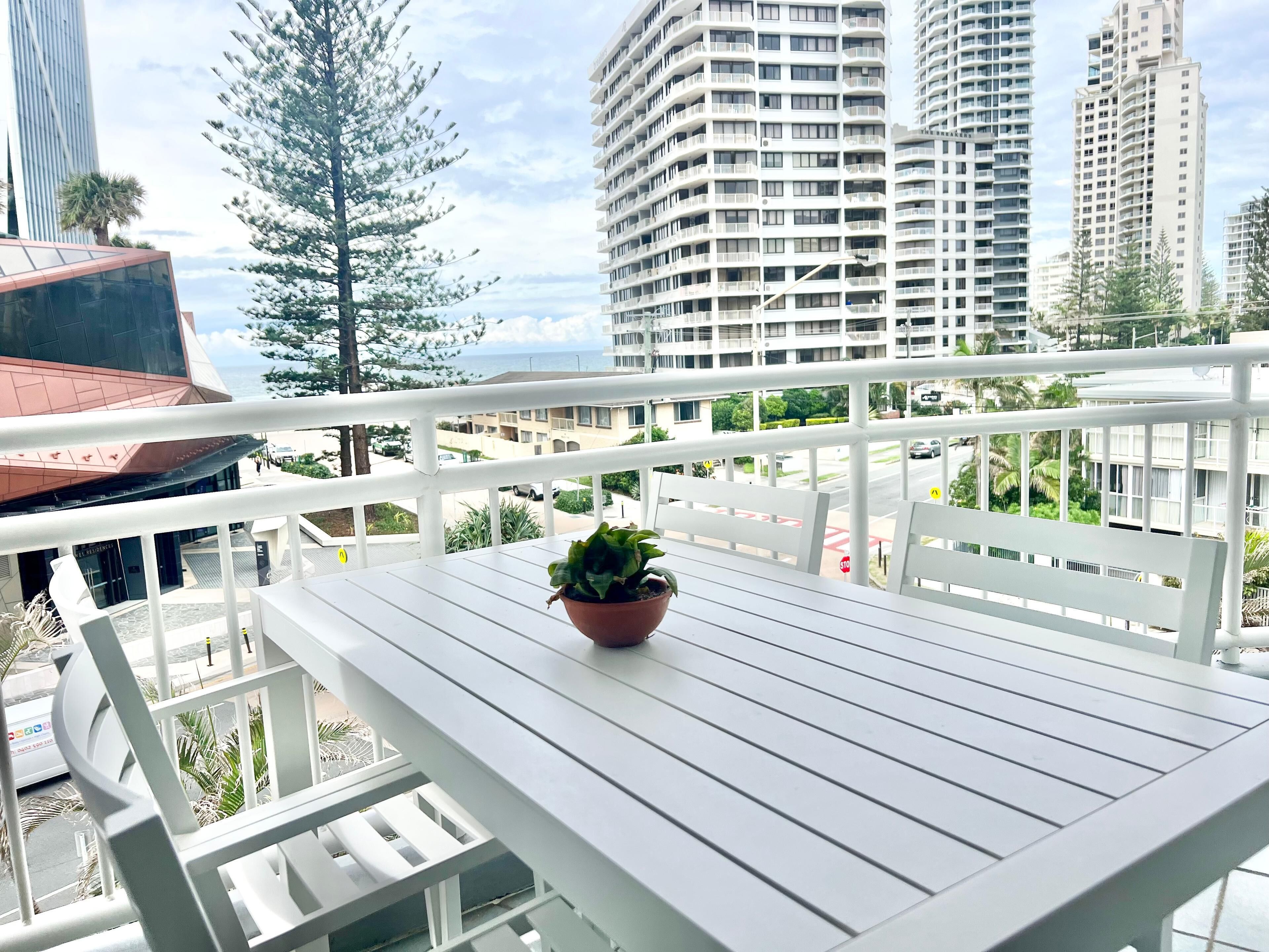 A balcony with a potted plant, white railings, and a view of high-rise buildings and palm trees.