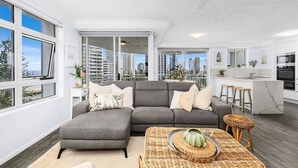 A modern apartment with a grey sectional sofa, a kitchen with a marble countertop, and large windows offering a city view. - Wharf Boutique Apartments (Surfers Paradise)