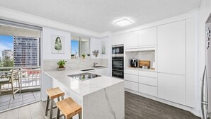 A modern kitchen with white cabinetry, a large island with stools, and a view of the city through the balcony. - Wharf Boutique Apartments (Surfers Paradise)
