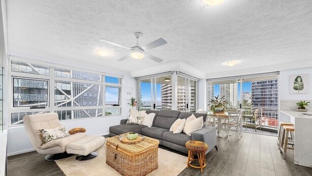 A spacious living area with a grey sofa, a beige armchair, and a wicker coffee table. There is a ceiling fan, large windows, and a balcony with a view of high-rise buildings. - Wharf Boutique Apartments (Surfers Paradise)