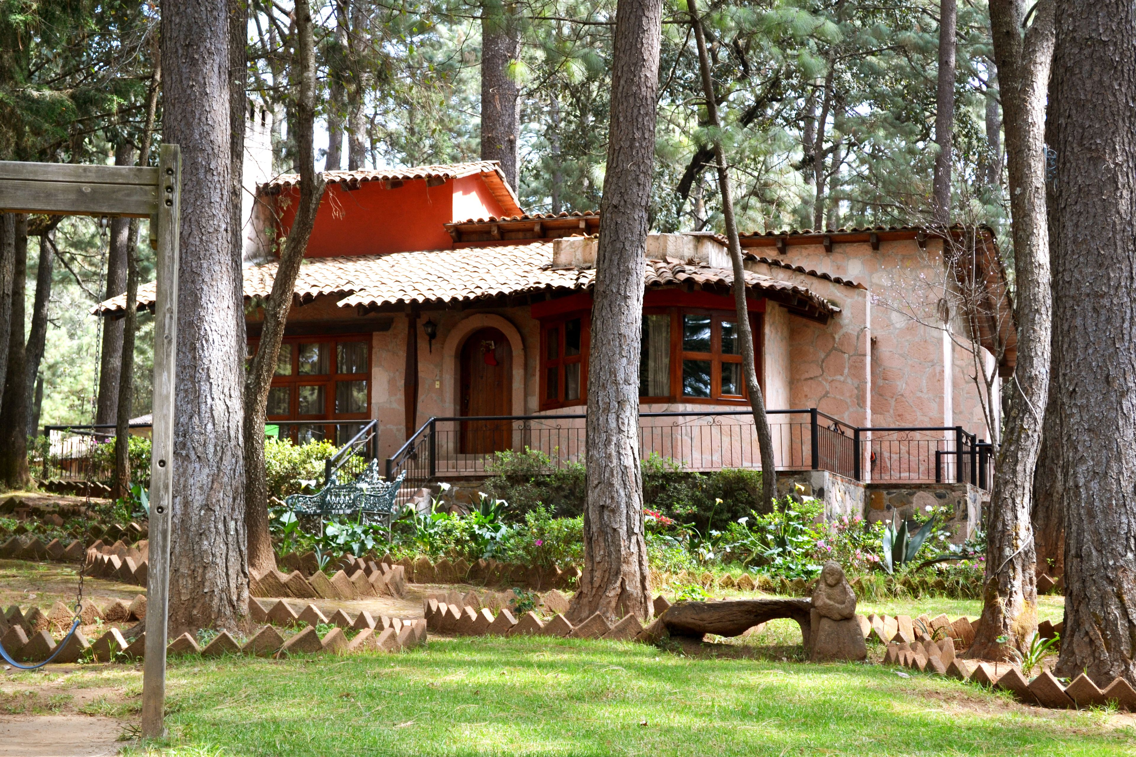 A rustic house with a red roof, surrounded by trees, and a small garden with a bench.