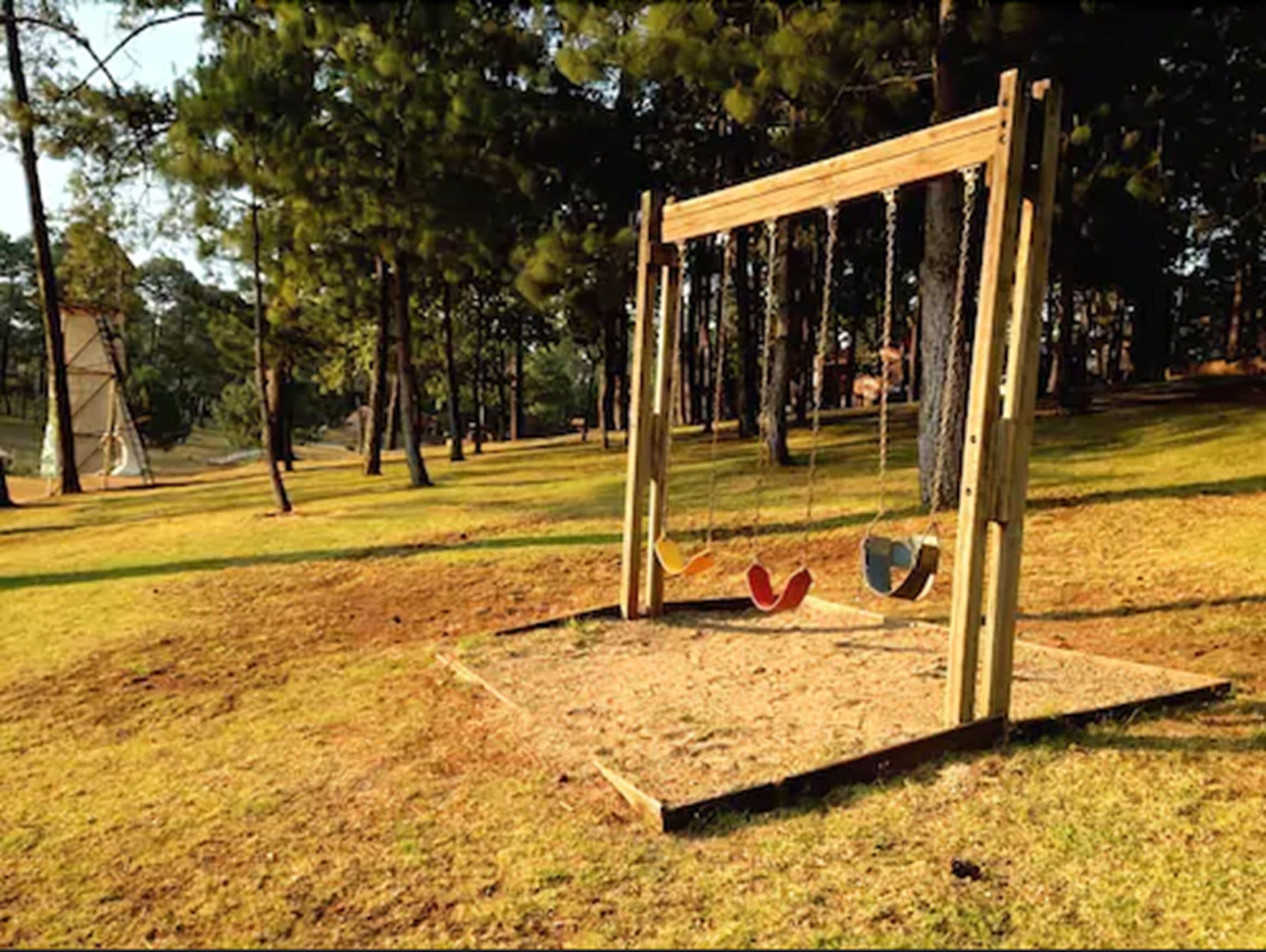 Un ensemble de balançoires en bois dans un parc, avec un sol sablonneux et des arbres en arrière-plan.