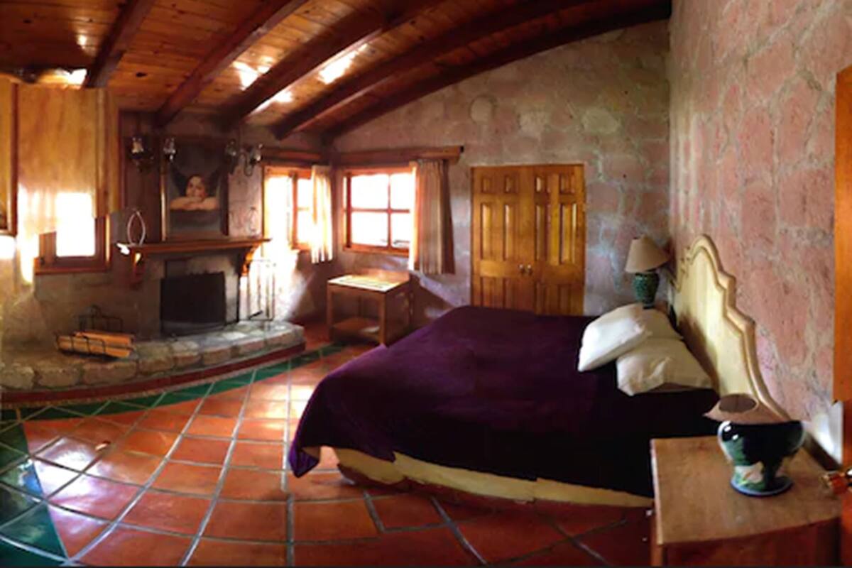 A rustic bedroom with a wooden ceiling, a stone fireplace, a bed with a purple bedspread, and a stone wall.