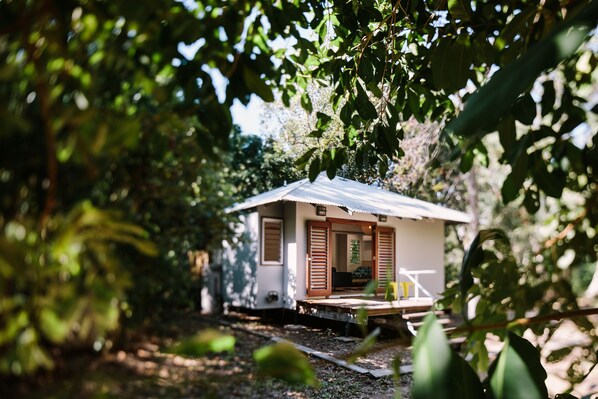 A small, single-story house with a flat roof, wooden shutters, and a porch. - The Little Bush Hut (Nelly Bay)
