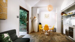 A modern kitchen with white cabinets, a marble countertop, and stainless steel appliances. There is a dining area with a yellow table and chairs, and a large window providing a view of greenery outside. - The Little Bush Hut (Nelly Bay)