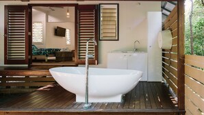 A modern bathroom with a freestanding bathtub, wooden deck, and a view of the living area. - The Little Bush Hut (Nelly Bay)